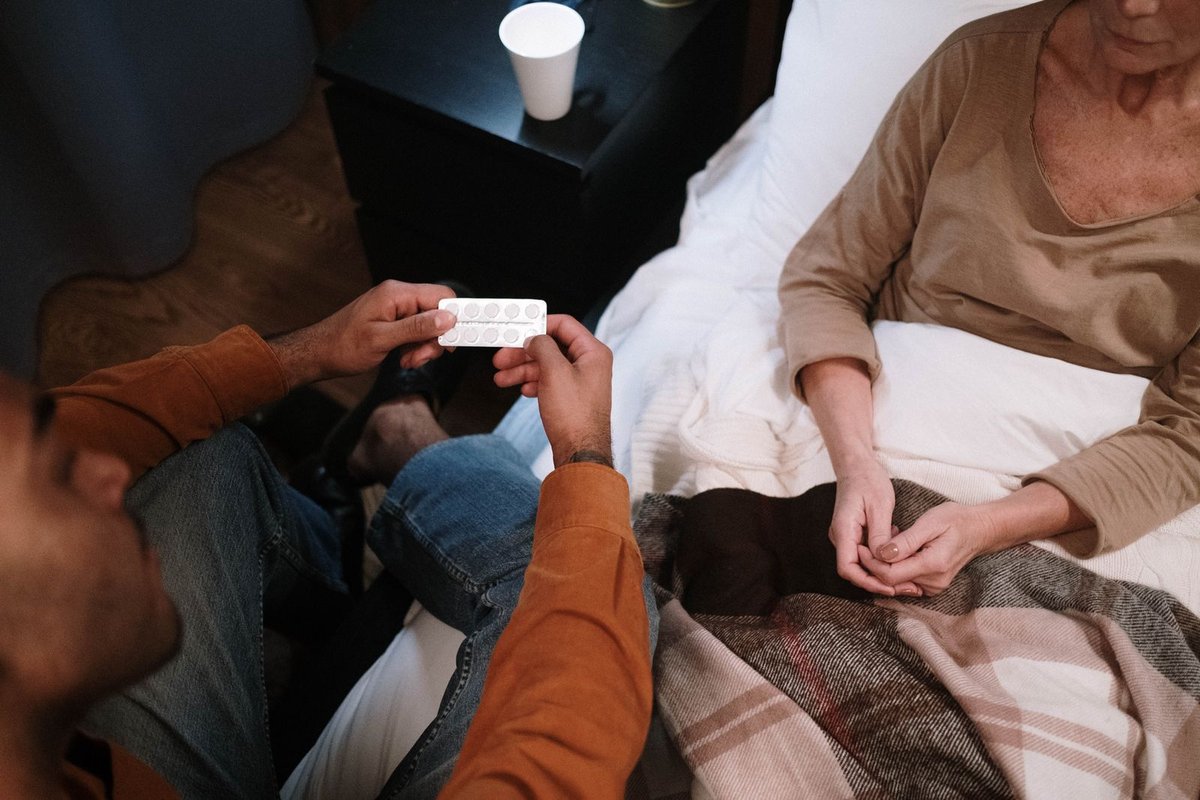 Caregiver sitting on a bed edge showing a blister pack of white pills to a senior under a plaid blanket