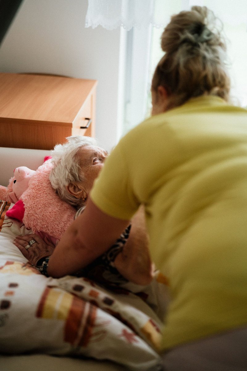 Caregiver in a yellow shirt leaning over an elderly woman with white hair lying in bed